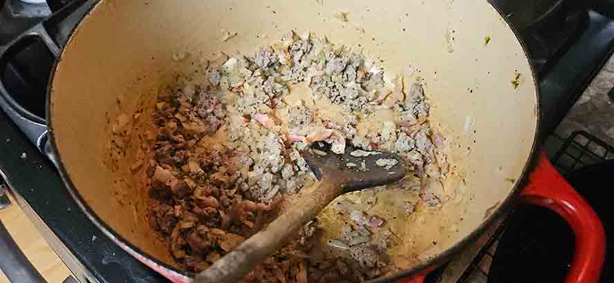 Browning crumbled sausages with onions and bacon in a thick-bottomed pot."