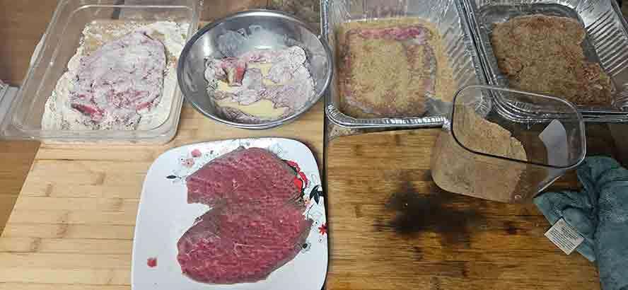A three-step breading station for beef schnitzel with separate bowls for flour, beaten eggs, and breadcrumbs."