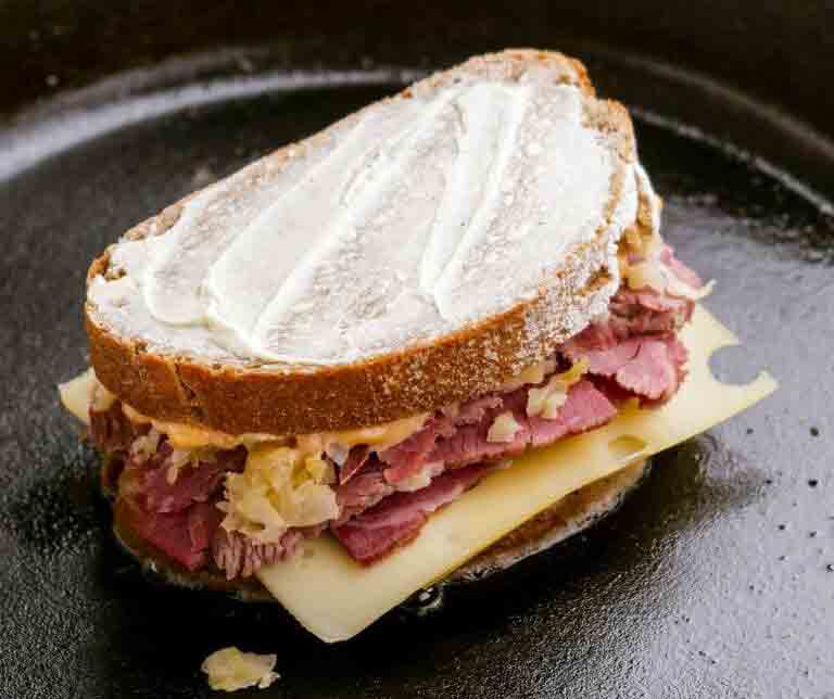 reuben-method-1 A Reuben sandwich with corned beef, melted Swiss cheese, and sauerkraut on marbled rye bread being grilled in a cast iron skillet."