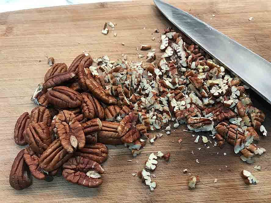 Pecans being chopped into small pieces on a wooden cutting board.